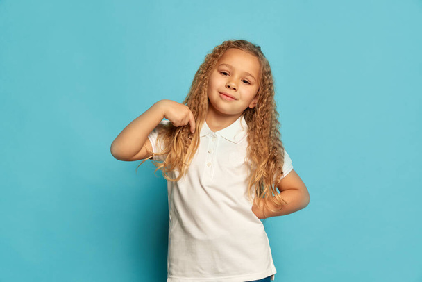Smiling child. Cute little happy girl, kid with long curly hair pointing at herself at camera isolated over blue background. Concept of children positive emotions, beauty, facial expressions. Copy - Foto, Bild