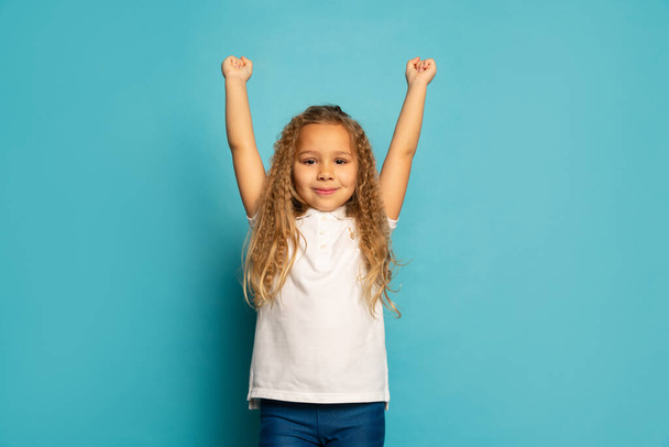 Win, winner. Cute little happy girl, kid with long curly hair looking at camera isolated over blue background. Concept of children positive emotions, beauty, facial expressions. Copy space for ad - Foto, imagen
