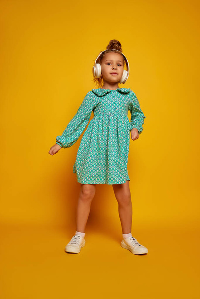 Dancing child. Cute little happy girl, kid with headphones looking at camera isolated over yellow background. Concept of children positive emotions, beauty, facial expressions. Copy space for ad - Φωτογραφία, εικόνα