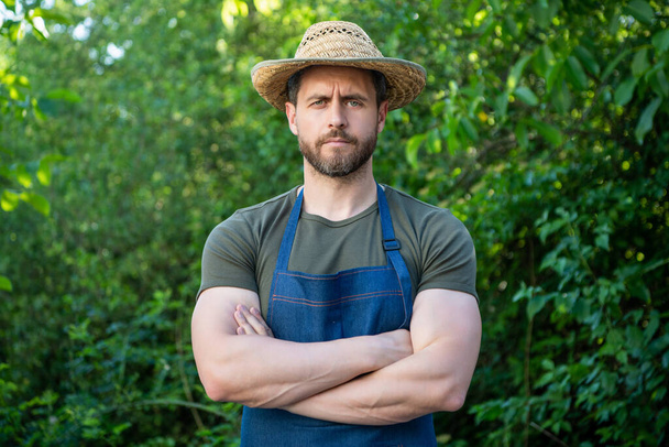 Confident man gardener in gardening apron and farmers hat keeping arms crossed standing in garden outdoors. - 写真・画像