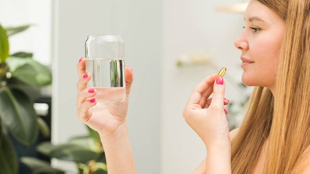 Happy blonde woman holding a glass of water and a capsule of fish oil or vitamin D in the kitchen. Vitamins and nutritional supplements. Healthy lifestyle. Preventive medicine. - Photo, image