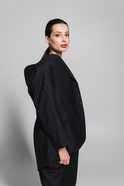Stylish brunette model in black suit looking at camera on grey background  - Foto, afbeelding