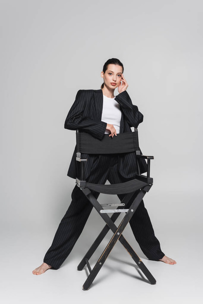 Longitud completa de la mujer de moda en traje posando cerca de la silla plegable sobre fondo gris  - Foto, imagen
