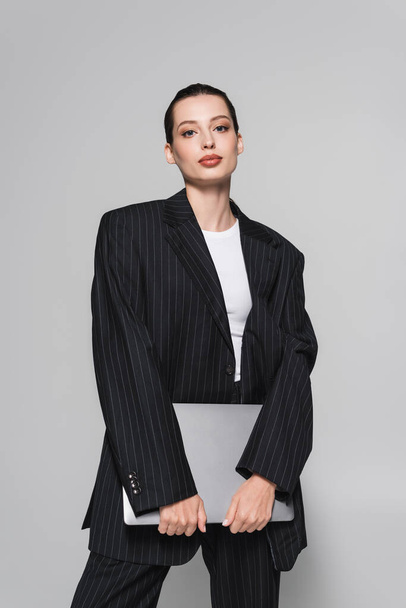 Fashionable woman in suit holding laptop and looking at camera isolated on grey  - Foto, immagini