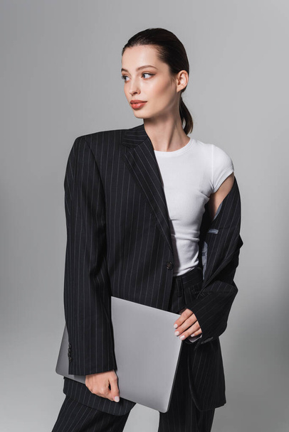 Pretty brunette woman in striped suit holding laptop isolated on grey  - Foto, Imagen