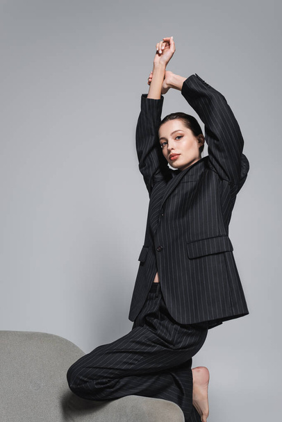 Barefoot model in striped suit raising hands near armchair isolated on grey  - Fotografie, Obrázek