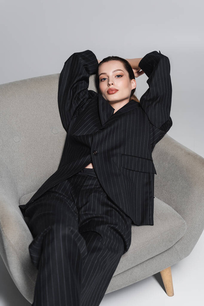 Trendy young woman in black striped suit looking at camera on armchair on grey background  - Foto, Bild