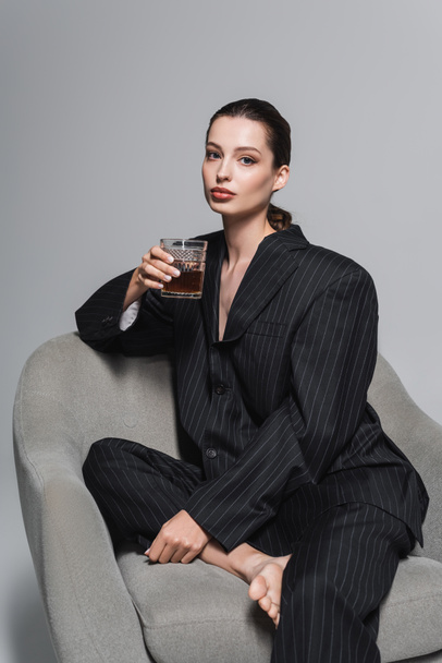 Stijlvol model in zwart pak met een glas whisky terwijl je op een fauteuil zit op een grijze achtergrond  - Foto, afbeelding