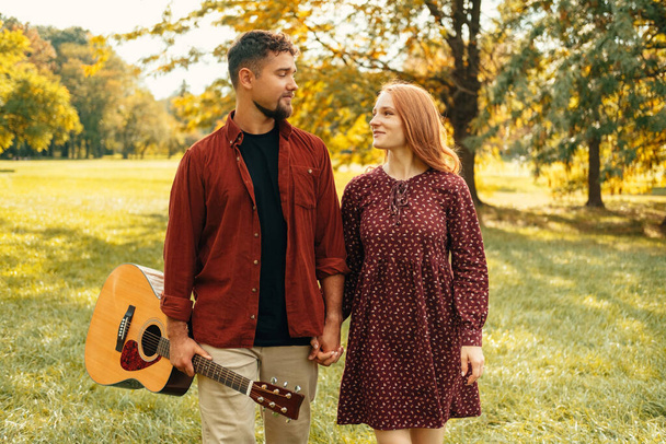 Młoda szczęśliwa para idzie na spacer w słonecznym zielonym parku z gitarą akustyczną w czerwonych ubraniach. - Zdjęcie, obraz
