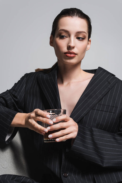 Πορτρέτο της γυναίκας σε ριγέ σακάκι κρατώντας ποτήρι ουίσκι, ενώ κάθεται στην πολυθρόνα απομονώνονται σε γκρι  - Φωτογραφία, εικόνα