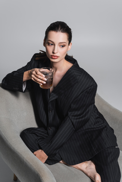 Brunette woman in striped suit holding whiskey while sitting on armchair on grey background  - Foto, afbeelding