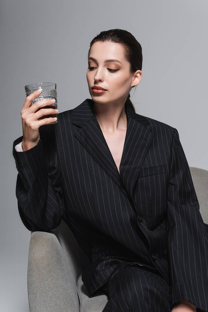 Stylish woman in striped jacket looking at glass of whiskey on armchair isolated on grey  - Foto, Imagen
