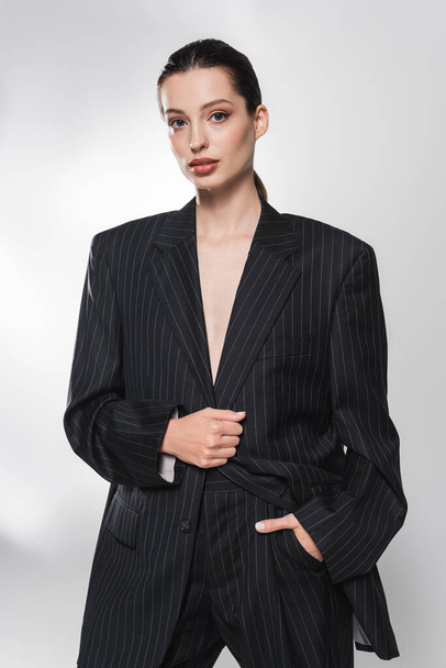 Портрет модной модели в костюме, держащей руку в кармане на сером фоне  - Фото, изображение