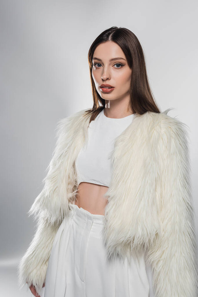 Stylish woman in white faux fur jacket looking at camera on grey background  - Photo, Image