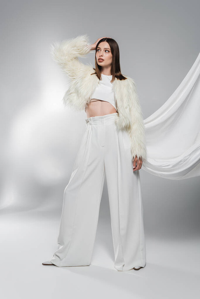 Trendy woman in faux fur jacket and white pants posing on abstract grey background  - Foto, afbeelding