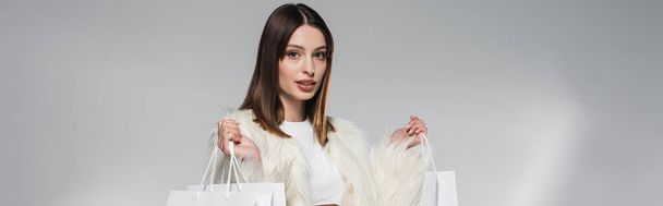 Stylish model in white faux fur jacket holding shopping bags on grey background, banner  - Fotografie, Obrázek
