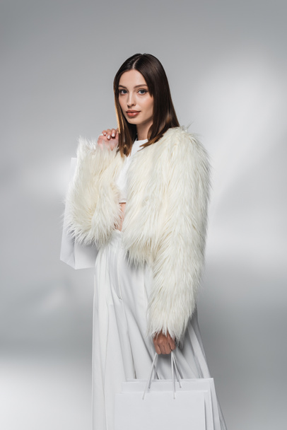 Trendy brunette woman in white faux fur jacket holding shopping bags on abstract grey background  - Photo, Image