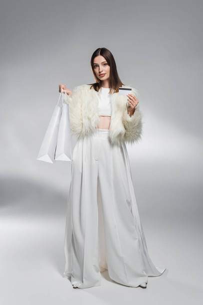 Full length of trendy woman in white clothes holding credit card and shopping bags on abstract grey background  - 写真・画像