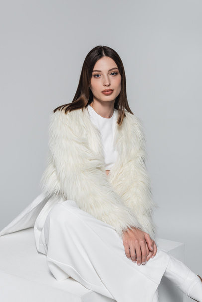young woman in white faux fur jacket and total white outfit sitting on cube isolated on grey - Foto, imagen