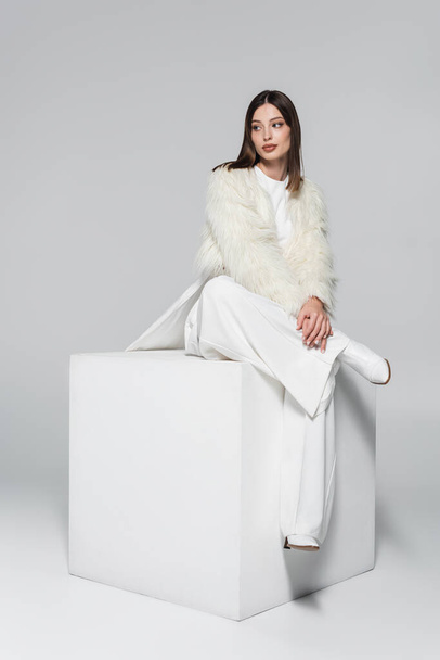 full length of young woman in faux fur jacket and total white outfit sitting on cube on grey - Valokuva, kuva