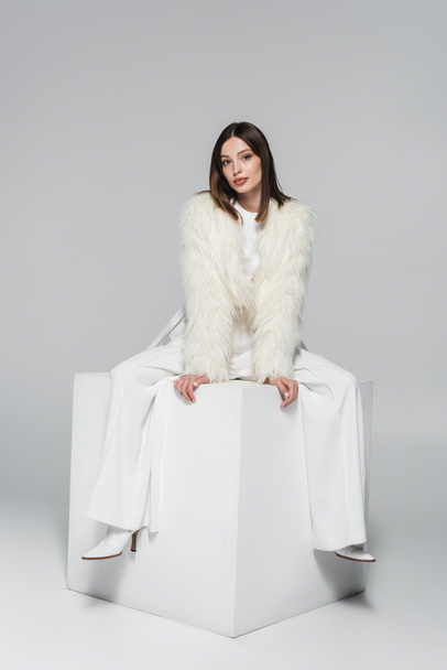 full length of stylish woman in faux fur jacket and total white outfit sitting on cube on grey - Valokuva, kuva