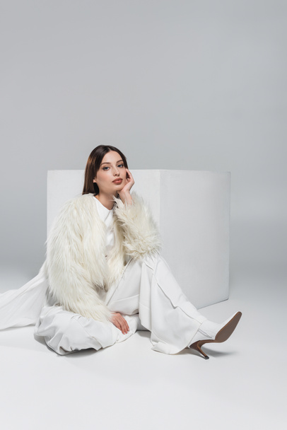 full length of young woman in faux fur jacket and total white outfit sitting near cube on grey - Φωτογραφία, εικόνα