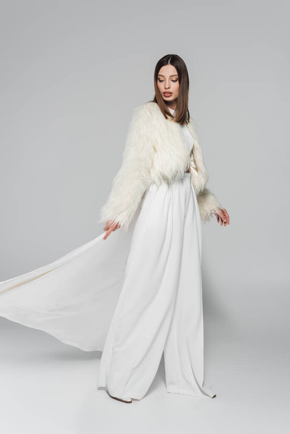 piena lunghezza di bella donna in abito totalmente bianco e giacca di pelliccia sintetica in posa sul grigio - Foto, immagini