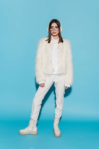 full length of stylish woman in earmuffs and eyeglasses posing in faux fur jacket on blue - Foto, afbeelding