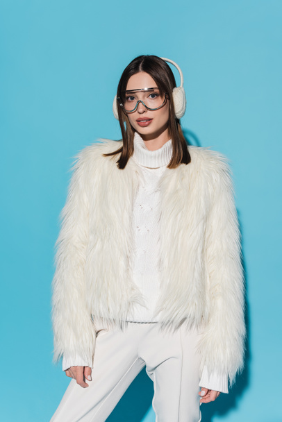 stylish young woman in earmuffs and eyeglasses posing in faux fur jacket on blue - Valokuva, kuva