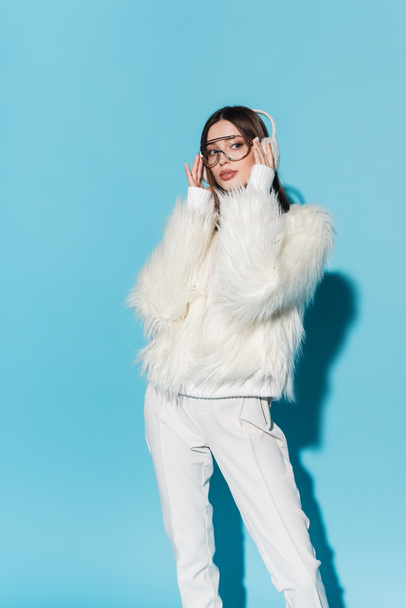 stylish young woman in earmuffs adjusting trendy eyeglasses while posing in faux fur jacket on blue - 写真・画像