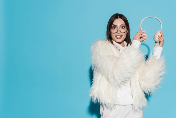 amazed young woman in eyeglasses and stylish faux fur jacket holding winter earmuffs on blue  - Foto, afbeelding