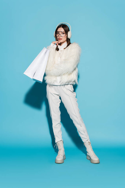 full length of stylish woman in faux fur jacket and eyeglasses holding shopping bags on blue - Photo, Image