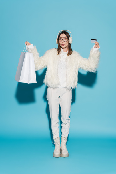 full length of surprised woman in eyeglasses and stylish faux fur jacket holding credit card and shopping bags on blue - Foto, Imagem