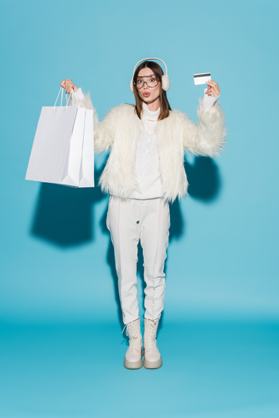 teljes hossza meglepett nő fülvédővel és fehér faux szőrme kabát gazdaság hitelkártya és bevásárló táskák kék - Fotó, kép