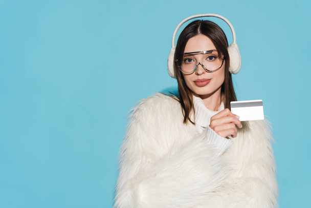 zufriedene junge Frau mit Brille und stylischer Kunstpelzjacke, Kreditkarte isoliert auf blauem Grund   - Foto, Bild