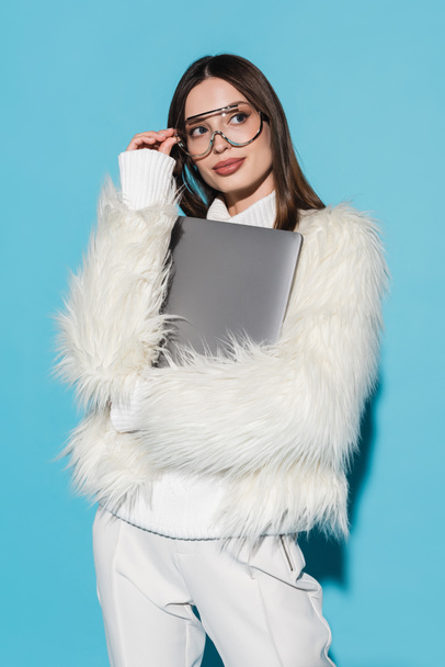 young woman in trendy eyeglasses and stylish faux fur jacket holding laptop on blue - Zdjęcie, obraz