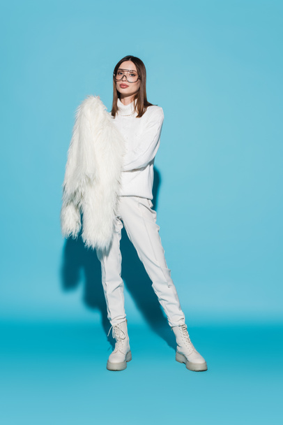 piena lunghezza di elegante giovane donna in occhiali alla moda in possesso di giacca di eco pelliccia bianca su blu - Foto, immagini