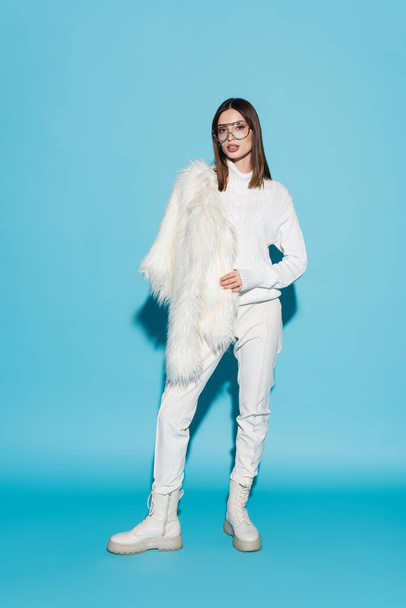 full length of stylish woman in trendy eyeglasses and white turtleneck holding white faux fur jacket on blue - Foto, immagini