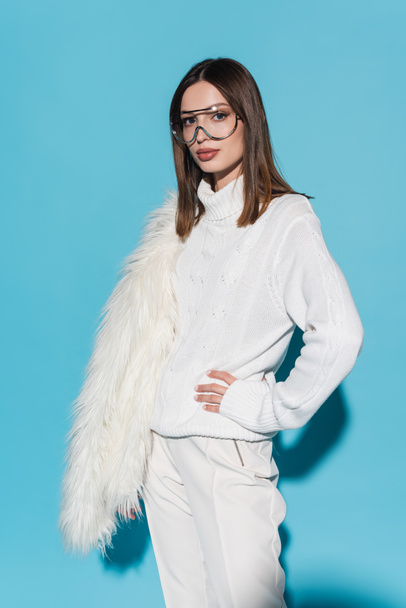 stylish woman in eyeglasses and white turtleneck standing with hand on hip while holding faux fur jacket on blue - Zdjęcie, obraz