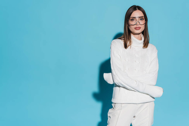 pretty young woman in trendy eyeglasses and white turtleneck posing with crossed arms on blue background - Photo, Image