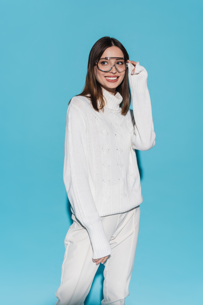 joyful young woman in trendy eyeglasses and totally white outfit posing on blue - Photo, Image