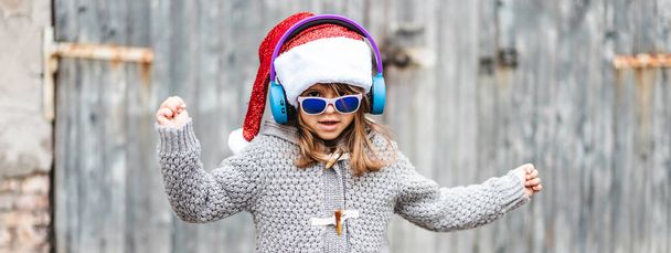Οριζόντια λάβαρο ή κεφαλιά με αστείο κοριτσάκι φορώντας καπέλο Αϊ Βασίλη και γυαλιά ηλίου χορεύουν σε ξύλινο φόντο σε εξωτερικούς χώρους τα Χριστούγεννα - Παιδική έννοια στις ημέρες των Χριστουγέννων - Φωτογραφία, εικόνα
