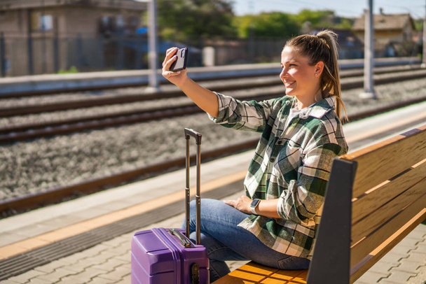 Happy woman is taking selfie with mobile phone while sitting on a train station. - Фото, изображение