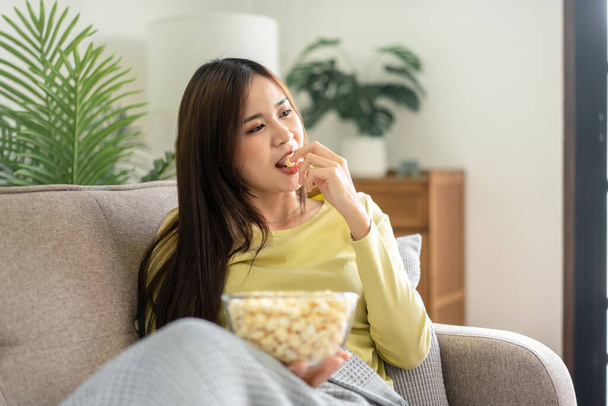 Joven mujer asiática sosteniendo un tazón de palomitas de maíz para comer palomitas de maíz mientras está acostado en el gran sofá cómodo para ver películas en la televisión en la sala de estar en casa. - Foto, Imagen