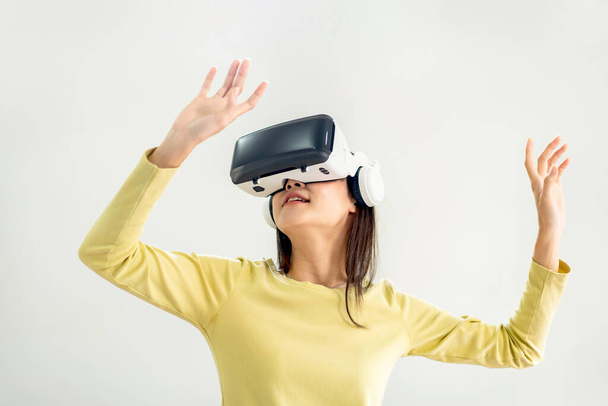 Młoda azjatycka kobieta w okularach wirtualnej rzeczywistości do oglądania wideo i grania w gry z wirtualnym doświadczeniem, używając rąk z palcami do dotykania w powietrzu i stojąc w salonie w domu. - Zdjęcie, obraz
