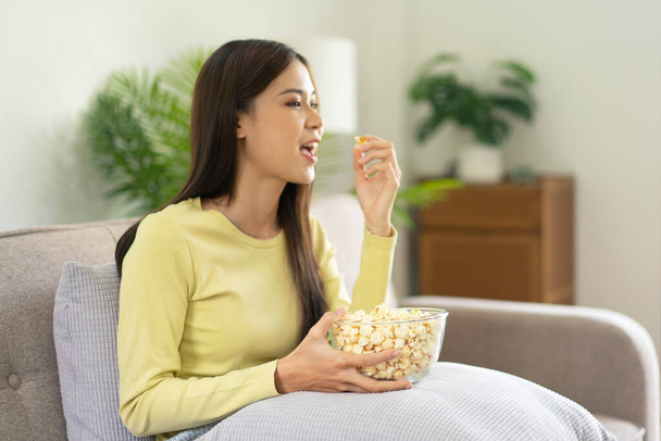 Giovane donna asiatica mettere una ciotola di popcorn sul cuscino e mangiare popcorn mentre seduto sul grande comodo divano a guardare film in televisione in soggiorno a casa. - Foto, immagini
