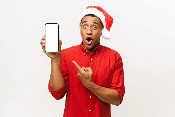 Здивований афроамериканець у капелюсі в Санті вказує на мобільний телефон з чистим екраном ізольованим на білому, хлопець у червоній сорочці задоволений повідомленням, дивлячись в камеру з відкритим ротом - Фото, зображення