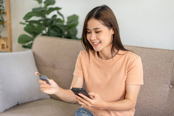 Young asian woman is holding new credit card and smartphone while sitting on comfortable sofa and entering information on banking for shopping online in living room at home. - Photo, Image