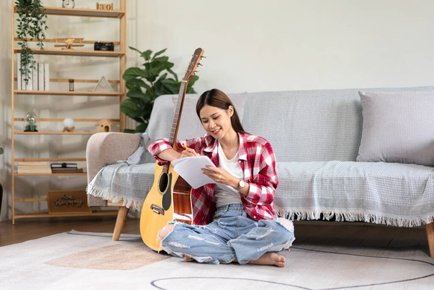 Genç bir kadın dizüstü bilgisayara şarkı besteleyip şarkı sözü yazarken bacaklarına gitar takıyor ve evdeki oturma odasında yere oturuyor.. - Fotoğraf, Görsel
