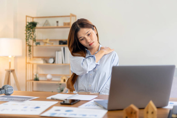 Vrouwelijke freelance voelt pijn in schouder en rug tijdens het lezen van business report op laptop en het werken op de tafel in de woonkamer op kantoor. - Foto, afbeelding